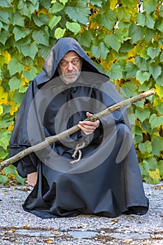 Severe Monk in a black robe with a staff on a background of grape leaves. Mysticism and magic,