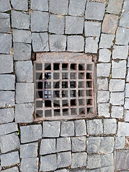 A sewer drain with an iron grate on a stone cobblestone sidewalk