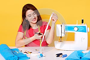 Sewer wears red casual t shirt with happy expression sitting with happy expression, cutting white ribbon, posing around sewing