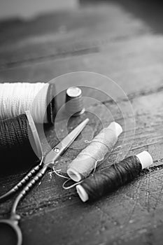 Sewing accessories: bobbins of thread, scissors, needle, thimble on wooden table. Black and white photo. Tailoring and