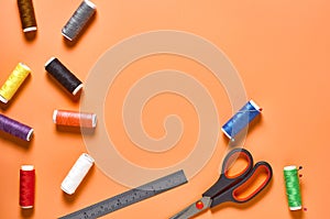 Sewing accessories scissors, metal ruler, spools with colorful threads and needles on orange tailors desk in workshop