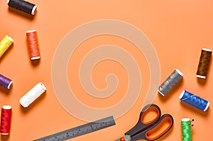 Sewing accessories scissors, metal ruler, spools with colorful threads and needles on orange tailors desk in workshop