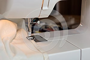 The sewing machine`s foot with a needle sews ecru color fabric. Sewing machine details