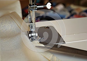 The sewing machine`s foot with a needle sews ecru color fabric. Sewing machine details