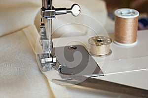 The sewing machine`s foot with a needle sews ecru color fabric. Sewing machine details