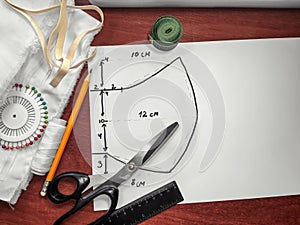 Sewing supplies on background of a drawn pattern of a medical face mask. Scissors, ruler, pencil, thread, fabric and white sheet