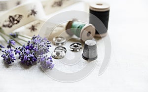 Sewing tools with fresh lavander flowers on linen background. Vintage wooden spool, braid, thimble, buttons.