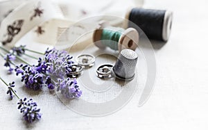 Sewing tools with fresh lavander flowers on linen background. Vintage wooden spool, braid, thimble, buttons.