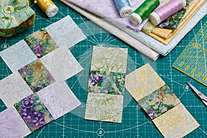 Sewn square pieces of fabric, a stack of fabrics, sewing and quilting accessories lying on a cutting mat