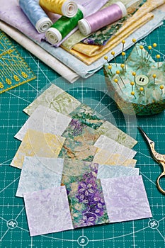 Sewn square pieces of fabric, a stack of fabrics, sewing and quilting accessories lying on a cutting mat