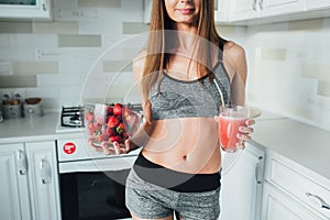 Sexual sports girl holding a bowl of strawberries and a glass of smoothie in the kitchen.