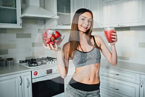 Sexual sports girl holding a bowl of strawberries and a glass of smoothie in the kitchen.