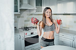 Sexual sports girl holding a bowl of strawberries and a glass of smoothie in the kitchen.