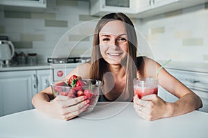 Sexual sports girl holding a bowl of strawberries and a glass of smoothie in the kitchen.