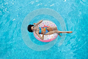 Sexy African American woman with summer drink relaxing on inflatable ring at swimming pool, overhead view