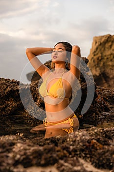 Sexy Asian woman posing on the beach. Beautiful woman wearing yellow bikini. Slim fit body. Tanned skin. Ocean water. Sunset time