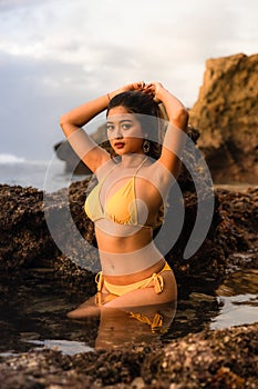 Sexy Asian woman posing on the beach. Beautiful woman wearing yellow bikini. Slim fit body. Tanned skin. Ocean water. Sunset time