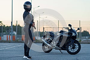 Sexy biker woman in black leather jacket and full face helmet stands near stylish sports motorcycle. Urban parking