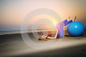 Sexy blonde with fitness ball on the beach outdoors. Beauty lies on the sand in the evening light against the sunset over the sea