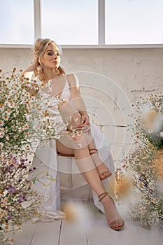 Sexy blonde woman in a beautiful white dress is sitting near the window in front of a bouquet of wild flowers. Romantic girl with