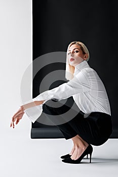 Sexy blonde woman with red lips wearing white shirt and black classic pants posing on black studio background
