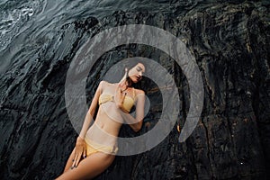 brunette in bikini lies on a large rock on ocean coast