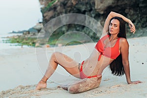 Sexy brunette girl in red swimsuit lies and sunbathes on beach with rocks.