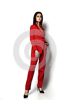 Sexy brunette woman in stylish red office pantsuit and high-heeled shoes is posing with her foot outstretched