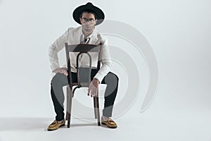 sexy cool man with hat and glasses sitting and holding elbow on knee