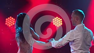 Sexy couple dancing salsa in a dark smoky studio with red lights in the background. A man in a white shirt is whirling