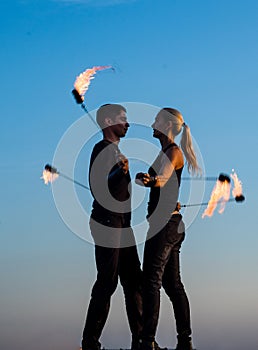 Sexy couple of fire dancers spin flaming poi on idyllic evening blue sky outdoors, motion