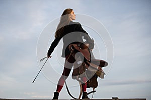 Sexy cowgirl hold saddle and whip. Equestrienne horsewoman. American cowboy style. Cowgirl woman in countryside