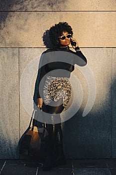 Sexy curvy afro woman is leaning against a wall and is on her phone looking down. She is holding her purse
