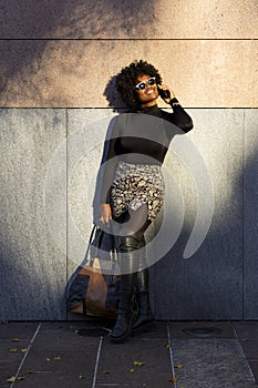 Sexy curvy afro woman is leaning against a wall and is on the phone smiling and looking up. She is holding her purse