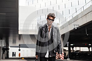 Sexy European young man in sunglasses in fashion youth oversized black leather jacket with a trendy hairstyle is resting near a