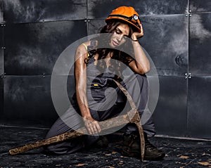 female miner worker with pickaxe, in coveralls over his naked body, sitting on the floor on backdrop of steel wall