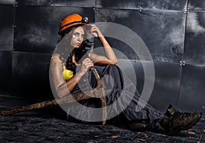 female miner worker with pickaxe, in coveralls over his naked body, sitting on the floor on backdrop of steel wall