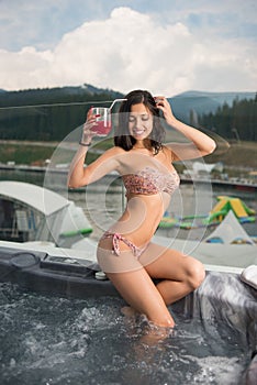 female with perfect figure in bikini sitting on the Jacuzzi lowered feet in the water and drinking cocktail