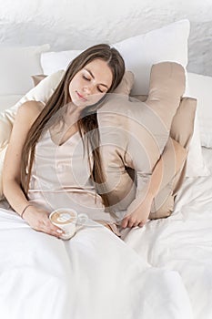 Sexy girl with a beauty pillow and coffee cup in her hands sits in bed. Vertical photo