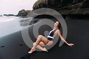 Sexy girl with big breasts in blue swimwear relaxing at beach with black sand.
