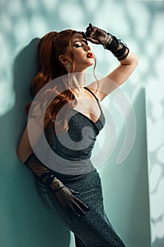 Sexy Girl in Black Dress leaning against Wall. Beautiful Red Hair Lady in Glitter Gown with Ponytail Hairstyle. Fashion Model side