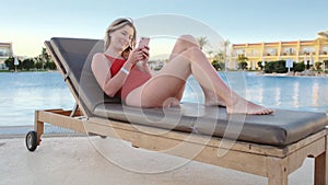 Sexy girl relaxing near the pool with a mobile phone. Woman texting and browsing the internet on mobile phone, lying on