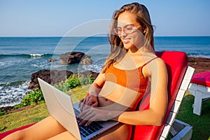 Sexy girl sitting on orange lounger with a laptop by the sea in the resort. beautiful business woman in stylish coral
