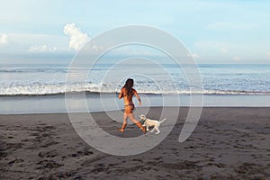 Sexy Girl. Tanned Woman Running On Sandy Beach. Beautiful Brunette In Bikini Playing With Dog