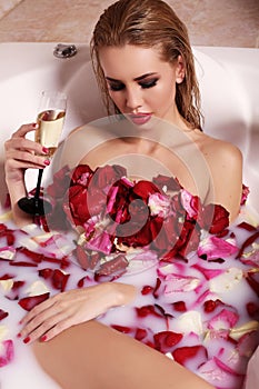 gorgeous woman with blond hair relaxing in bathroom with rose petals, drinking champagne