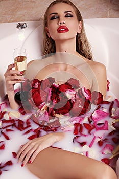 gorgeous woman with blond hair relaxing in bathroom with rose petals, drinking champagne