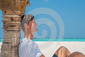 Sexy handsome model man in stylish clothes relaxing under palm tree at tropical sandy beach at island luxury resort