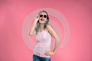 Sexy happy girl in sun glasses kiss shaped wearing tank top and denim jeans posing in studio isolated on pink background