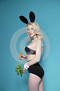 Sexy hot young blonde-haired model dressed in black lingerie with bunny ears, holding a carrot in her hand