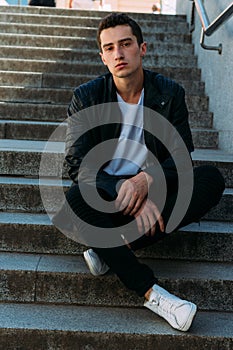 Sexy man posing sits on the steps near railing. Handsome young man in stylish black clothes and white shoes posing near a railing